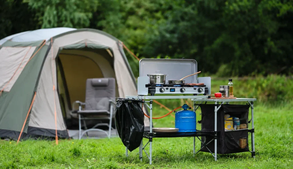 Two-burner gas camping stove on an outdoor cooking table beside a tent